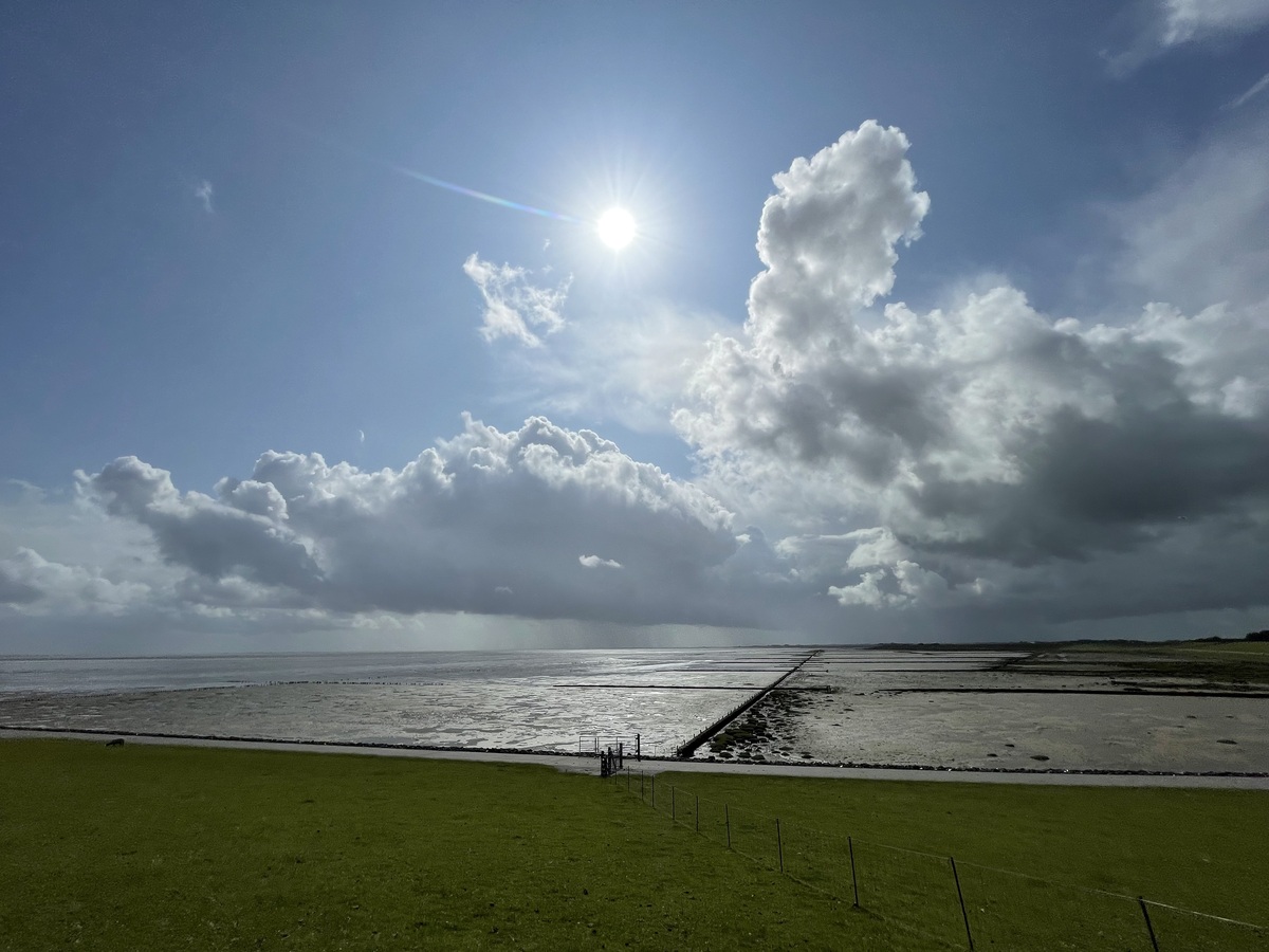 Der saftig grüne Deich und die Nordsee in Vollerwiek, am Himmel ein Wolken-Sonnen-Mix